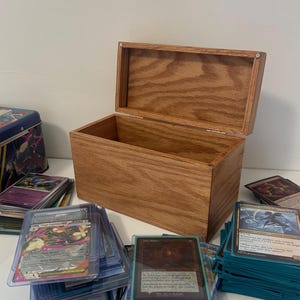 May include: A wooden card storage box with an open lid, showcasing its interior. The box is made of light brown wood and is surrounded by stacks of trading cards in protective sleeves. A blue tin is visible in the background.