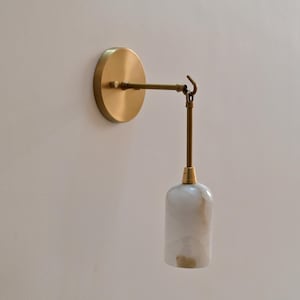 Könnte beinhalten: Eine Wandleuchte mit Messing-Finish. Sie verfügt über eine kreisförmige Basis, einen verstellbaren Arm und einen weiß-beige marmorierten Schirm. Das Design ist minimalistisch und modern, geeignet für die Innenbeleuchtung.