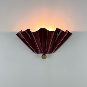 May include: A wall sconce featuring a deep burgundy, pleated, fan-shaped glass shade with a glossy finish. The light fitting is mounted with a small, centred, brushed brass spherical knob at the base, emitting a warm, soft glow from within.