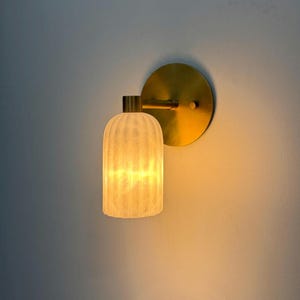 May include: A wall sconce featuring a circular brass backplate and a cylindrical, vertically ribbed frosted glass shade. The fixture emits a warm, soft glow from within the textured glass, mounted on a short horizontal brass arm.