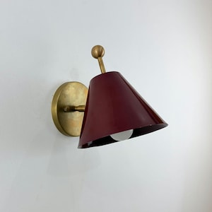 May include: A burgundy wall sconce with a conical shade and a brass-colored base. The sconce has a small, round light bulb and a brass-colored stem. The sconce is mounted on a white wall.