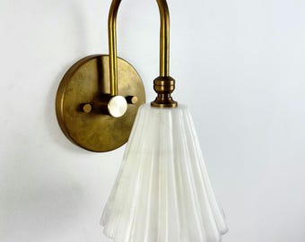 Op de afbeelding: Een wandlamp met een ronde messing muurplaat en een gebogen messing arm die een naar beneden gerichte, geribbelde, doorschijnende witte glazen kap ondersteunt. De metalen onderdelen hebben overal een geborstelde messing afwerking.