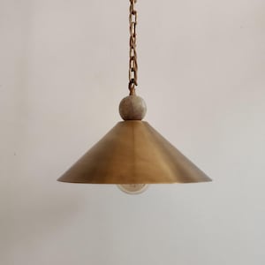May include: A brass pendant light with a conical shade and a decorative chain. The light hangs from a chain and features a small, round, stone-like detail above the shade. The light bulb is visible.