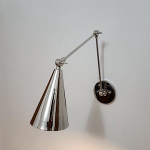 May include: A polished chrome wall lamp with a conical shade and adjustable arm. The lamp has a circular base and a reflective, metallic finish. The design is modern and minimalist, suitable for various interior styles.