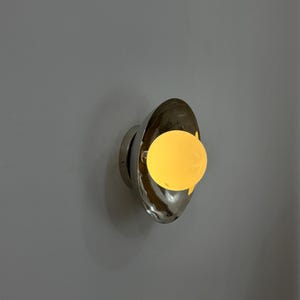 May include: A contemporary wall light with a polished chrome finish. The light has an oval design with a warm, yellow-toned bulb. The fixture is mounted on a grey wall, creating a minimalist aesthetic. A stylish addition to any home.