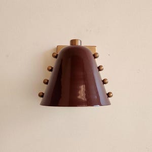May include: A wall-mounted lamp featuring a glossy, dark brown, bell-shaped shade. The shade is accented with three small, spherical, metallic-toned studs arranged vertically along each side. A metallic-toned base plate connects the lamp to the wall.