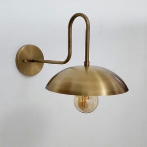 May include: A wall-mounted lamp featuring a brushed brass finish, a circular wall plate, and a curved arm that supports a wide, dome-shaped shade. A single exposed amber-tinted glass light bulb hangs downward from the center of the metal shade.