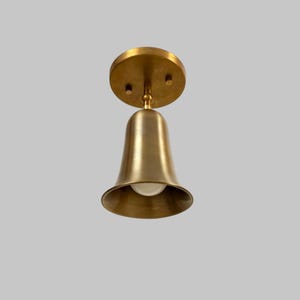 Peut inclure: Un luminaire de plafond en forme de cloche, de couleur laiton. Le luminaire a une base ronde avec trois trous de vis et un abat-jour en forme de cloche orienté vers le bas. Une seule ampoule est visible à l'intérieur de l'abat-jour. Le luminaire est conçu pour une utilisation en intérieur.