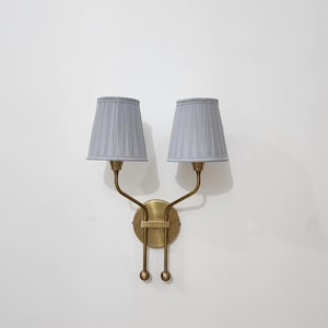 May include: A double wall light with two grey pleated lampshades. The fixture has a brass finish, with curved arms supporting the shades and a circular base. The design is symmetrical and elegant, suitable for interior lighting.