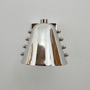 Op de afbeelding: Een gepolijste zilverkleurige wandlamp met een klokvormige kap en een glad, reflecterend oppervlak. Het armatuur is voorzien van vier kleine, bolvormige metalen noppen die verticaal aan weerszijden van de taps toelopende kap zijn geplaatst.
