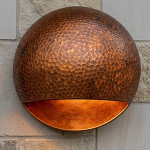 May include: A copper-toned, semi-circular wall light with a textured, hammered surface. The lower section has a smooth, polished finish, creating a contrast. The light is mounted on a light-coloured stone wall.