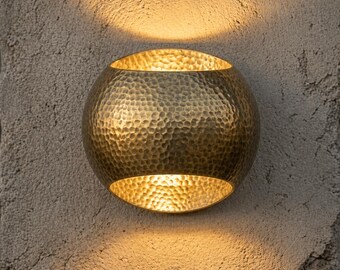 May include: A hammered brass wall sconce with a spherical shape. The sconce has an open top and bottom, emitting warm light. The fixture is mounted on a textured, light-colored wall, creating a contrast between the smooth metal and the rough surface.