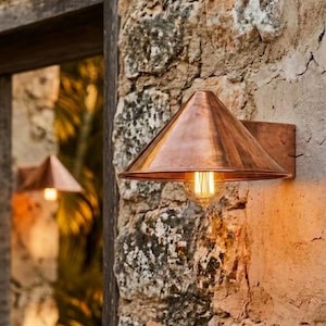 May include: A copper wall-mounted outdoor light fixture with a conical shade and a visible bulb. The sconce is fixed to a weathered stone wall, creating a rustic and warm atmosphere. The light fixture is a decorative element for exterior spaces.