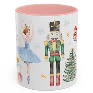 May include: White ceramic mug with a pink interior. It features watercolour illustrations of a ballerina, nutcracker, Christmas tree, and snowflakes. The mug has a glossy finish and is suitable for hot or cold drinks.