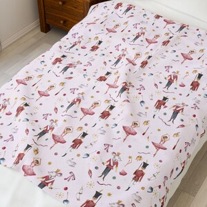 May include: A light pink blanket with a repeating pattern of watercolor illustrations. The design features ballerinas in pink tutus, toy soldiers in red coats, and mice in regal attire. Decorative elements include stars and ornaments.