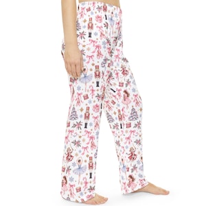 May include: White pajama pants featuring a repeating pattern of Nutcracker figures, ballet dancers, pink ribbons, Christmas trees, and snowflakes. The design includes small drums and candy canes scattered across the fabric in shades of pink and blue.