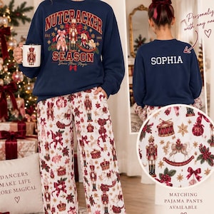 May include: A navy blue crewneck sweatshirt featuring a Nutcracker graphic on the front and the name Sophia printed on the back. The set includes white pajama pants with a repeating pattern of nutcrackers, drums, bows, and ballet slippers.