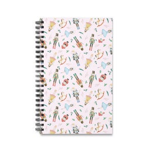 May include: Spiral-bound notebook with a pink cover featuring a repeating pattern of watercolor illustrations. The design includes nutcrackers, ballerinas, rocking horses, and perfume bottles. The notebook has a black spiral binding.