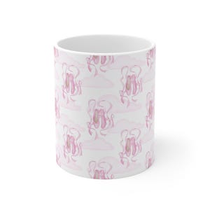 May include: White ceramic mug featuring a repeating pattern of pink ballet shoes, ribbons, and clouds. The design has a watercolor aesthetic, with soft pink hues and delicate details. The mug is cylindrical and has a white interior.