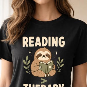 Può includere: T-shirt nera con la scritta "READING THERAPY" in crema. Un bradipo dei cartoni animati legge un libro con un cuore sulla copertina, circondato da foglie verdi e stelle. Design casual.