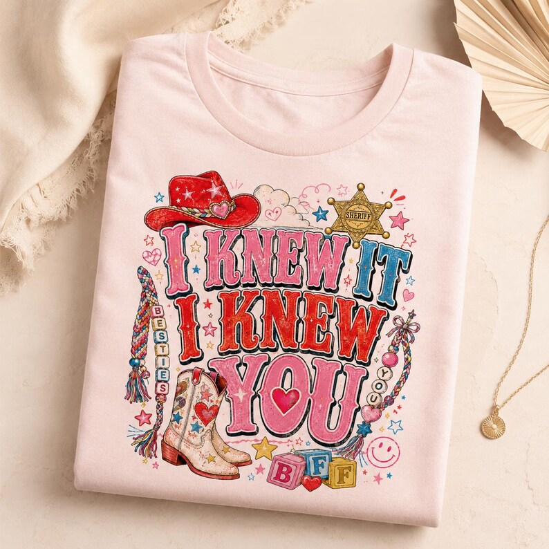 May include: A light pink t-shirt featuring a graphic print with the text I KNEW IT I KNEW YOU. The design includes a red cowboy hat, a gold sheriff badge, a cowboy boot, colorful braided friendship bracelets, and alphabet blocks spelling BFF.