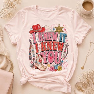 May include: A light pink short-sleeve t-shirt featuring a graphic print with the text I KNEW IT I KNEW YOU. The design includes a red cowboy hat, a sheriff badge, a cowboy boot, colorful braided strings, and alphabet blocks spelling BFF.