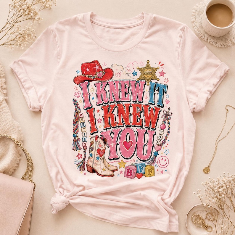 May include: A light pink short-sleeve t-shirt featuring a graphic print with the text I KNEW IT I KNEW YOU. The design includes a red cowboy hat, a sheriff badge, a cowboy boot, colorful braided strings, and alphabet blocks spelling BFF.