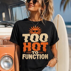 Op de afbeelding: Zwart T-shirt met de tekst "TOO HOT TO FUNCTION" in oranje en beige, met een zonnegrafiek met zonnebril. Het shirt wordt gedragen met een spijkerbroek, met een surfboard en een vintage oranje busje op de achtergrond.