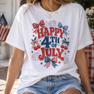 May include: A white short-sleeved t-shirt featuring a red, white, and blue graphic print that reads Happy 4th of July. The design includes illustrations of cherries, various patterned bows, and fireworks, all arranged in a festive, patriotic style.