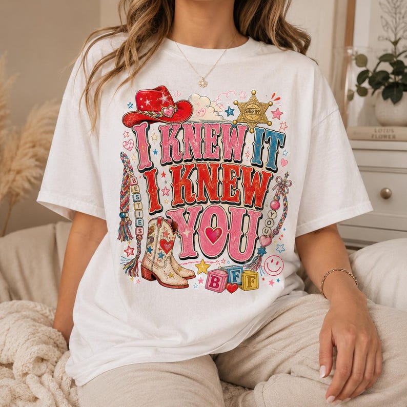 May include: A white short-sleeve t-shirt featuring a colorful graphic print with the text I KNEW IT I KNEW YOU. The design includes a red cowboy hat, a sheriff star, a cowboy boot, braided friendship bracelets, and alphabet blocks spelling BFF.
