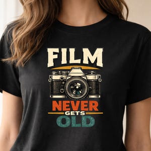 Op de afbeelding: Zwart T-shirt met een vintage camera-afbeelding en de tekst "FILM NEVER GETS OLD" in crème, oranje en teal. Het ontwerp toont een gedetailleerde illustratie van een klassieke filmcamera.