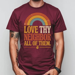 May include: Burgundy t-shirt with a retro rainbow design and the text "LOVE THY NEIGHBOR ALL OF THEM." The design includes a peace sign. The shirt is a crew neck style.