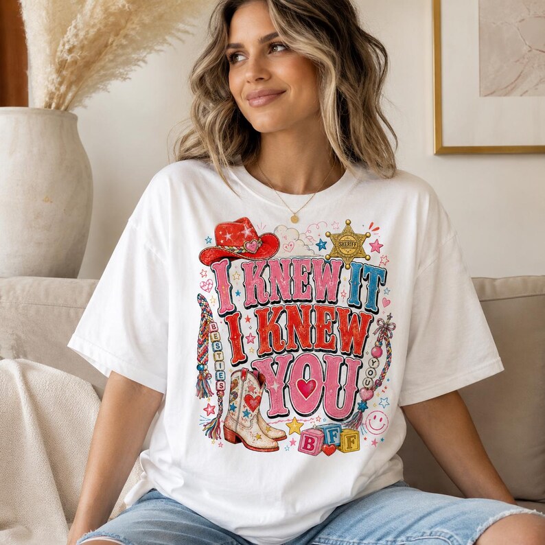 May include: A white t-shirt featuring a colorful graphic print with the text I KNEW IT I KNEW YOU. The design includes a red cowboy hat, a sheriff star, a cowboy boot, beaded friendship bracelets, and alphabet blocks spelling BFF.