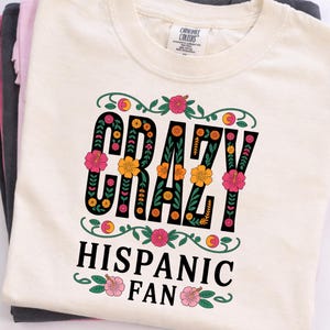 May include: Cream-colored t-shirt with the words "CRAZY HISPANIC FAN" in a floral design. The text is black with orange and pink flowers. The shirt is folded with other shirts in shades of gray and pink.
