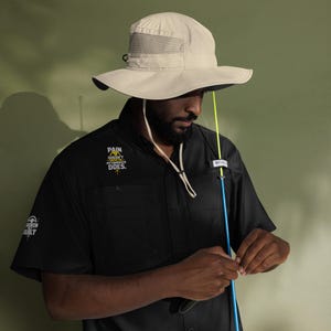 May include: A beige sun hat with a wide brim and mesh ventilation is paired with a black short-sleeve shirt. The shirt features the text "PAIN DOESN'T MY COMEBACK DOES." and "BROKEN BUILT." A fishing rod is held by the person.