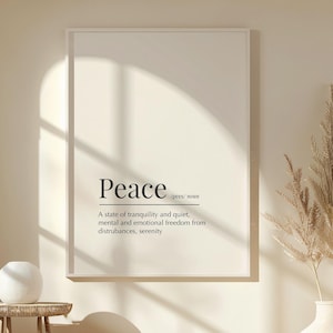 May include: A framed print with the word "Peace" and its definition in black text. The print hangs on a cream wall, with a white vase holding dried plants to the right. A small side table with a white sphere and a round ottoman are in the foreground.