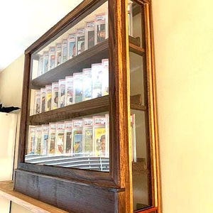May include: A wooden display case with glass panels and multiple shelves. The case is filled with graded cards. The frame is made of dark and light wood. The case is sitting on a wooden shelf.