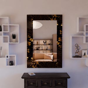 Pode incluir: Um espelho retangular com uma moldura decorativa preta e dourada está pendurado em uma parede branca. O espelho reflete uma sala com um sofá bege e prateleiras decorativas. Abaixo do espelho, há um armário de madeira marrom escuro.