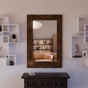 Op de afbeelding: Een rechthoekige spiegel met een decoratief bruin frame hangt boven een donkerhouten kast. De spiegel weerspiegelt een kamer met een bank en planken. Witte vierkante planken en decoraties bevinden zich aan de muur links en rechts van de spiegel.