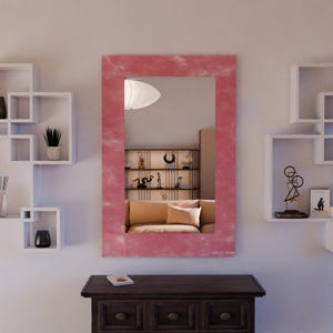 Könnte beinhalten: Ein rechteckiger Wandspiegel mit rosa Rahmen. Der Spiegel reflektiert einen Raum mit Sofa, Regalen und dekorativen Gegenständen. Der Spiegel ist über einem dunkelbraunen Holzschrank montiert.