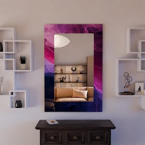 Puede incluir: Espejo de pared rectangular con un diseño abstracto vibrante en tonos morados, rosas y azules. El espejo enmarcado refleja una habitación con un sofá, estanterías y objetos decorativos. Está montado en una pared blanca sobre un mueble de madera oscura.