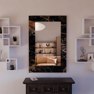 Puede incluir: Un espejo de pared rectangular con un marco con diseño de mármol negro. El espejo refleja una habitación con un sofá beige, estanterías y objetos decorativos. El espejo está montado sobre un mueble de madera oscura.