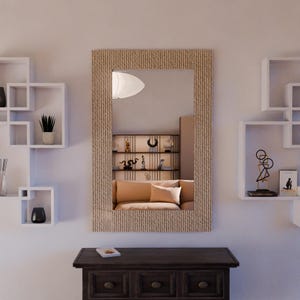 Può includere: Uno specchio rettangolare con una cornice strutturata marrone chiaro è appeso a una parete bianca. Un mobile in legno marrone scuro si trova sotto lo specchio. Mensole bianche sospese con oggetti decorativi su entrambi i lati.