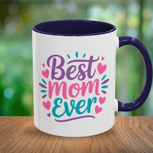 Puede incluir: Taza de cerámica blanca con interior y asa azul marino. La taza presenta el texto "Best Mom Ever" en morado, rosa y turquesa, con detalles de corazón y hojas. La taza está sobre una superficie de madera.