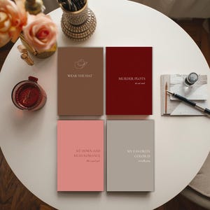 Peut inclure: Quatre cahiers à couverture rigide avec du texte sur une table blanche. Les cahiers sont marron, bordeaux, rose et gris. Le texte sur les cahiers se lit : "WEAR THE HAT", "MURDER PLOTS", "SIT DOWN AND READ ROMANCE", et "MY FAVORITE COLOR IS".