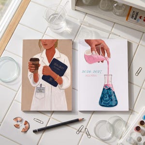Può includere: Due agende rettangolari con la scritta 2026-2027 sulle copertine. Una copertina raffigura una persona in camice bianco da laboratorio che tiene una tazza di caffè e un'agenda blu scuro. L'altra mostra una mano che versa liquido rosa in un matraccio.