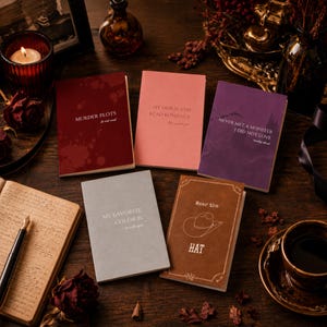 Peut inclure: Cinq carnets rectangulaires aux couvertures variées : un rouge foncé marqué MURDER PLOTS, un rose marqué SIT DOWN AND READ ROMANCE, un violet marqué NEVER MET A MONSTER I DID NOT LOVE, un gris marqué MY FAVORITE COLOR IS et un marron marqué WEAR THE HAT.