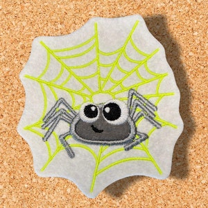 Könnte beinhalten: Ein gestickter Aufnäher mit einer grau-schwarzen Spinne mit einem lächelnden Gesicht auf einer unregelmäßig geformten weißen Form. Die Spinne sitzt auf einem neongrünen Netz. Der Aufnäher befindet sich auf einem Korkbrett-Hintergrund. Ein Halloween-Themen-Bastelartikel.