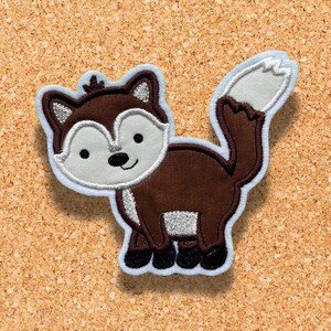 May include: A brown and white embroidered fox applique with a white tail. The fox is standing on a corkboard background.