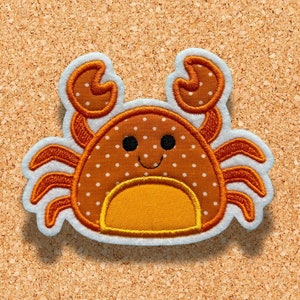 Könnte beinhalten: Ein gestickter Krabben-Patch in Braun und Orange mit weißen Punkten und einem lächelnden Gesicht. Der Krabbe befindet sich auf einem Korkuntergrund.